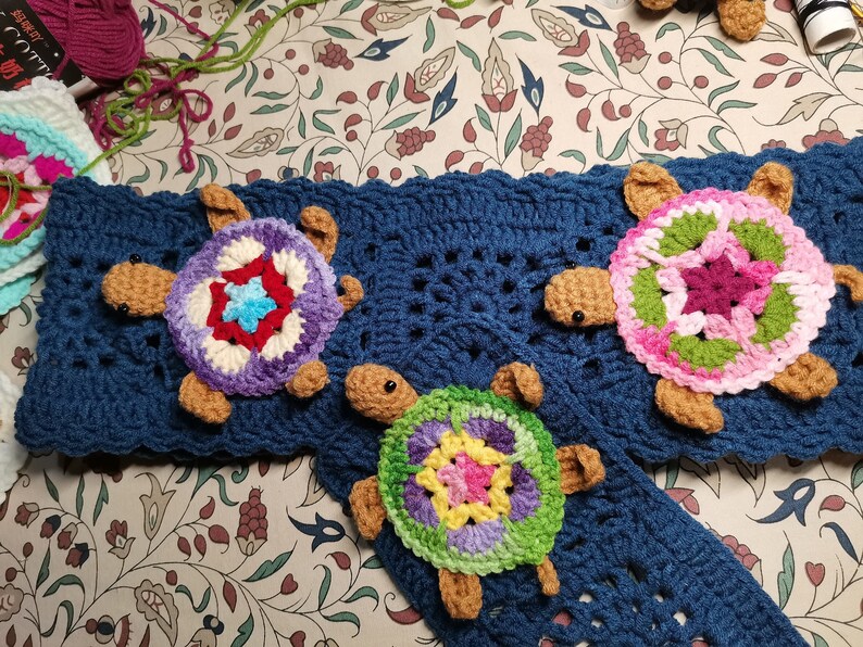 May include: Three crocheted turtle ornaments with colorful shell designs. The turtles are attached to a blue crocheted blanket.