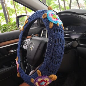 May include: A blue crocheted steering wheel cover with colorful crocheted turtle embellishments. The cover is on a car steering wheel.