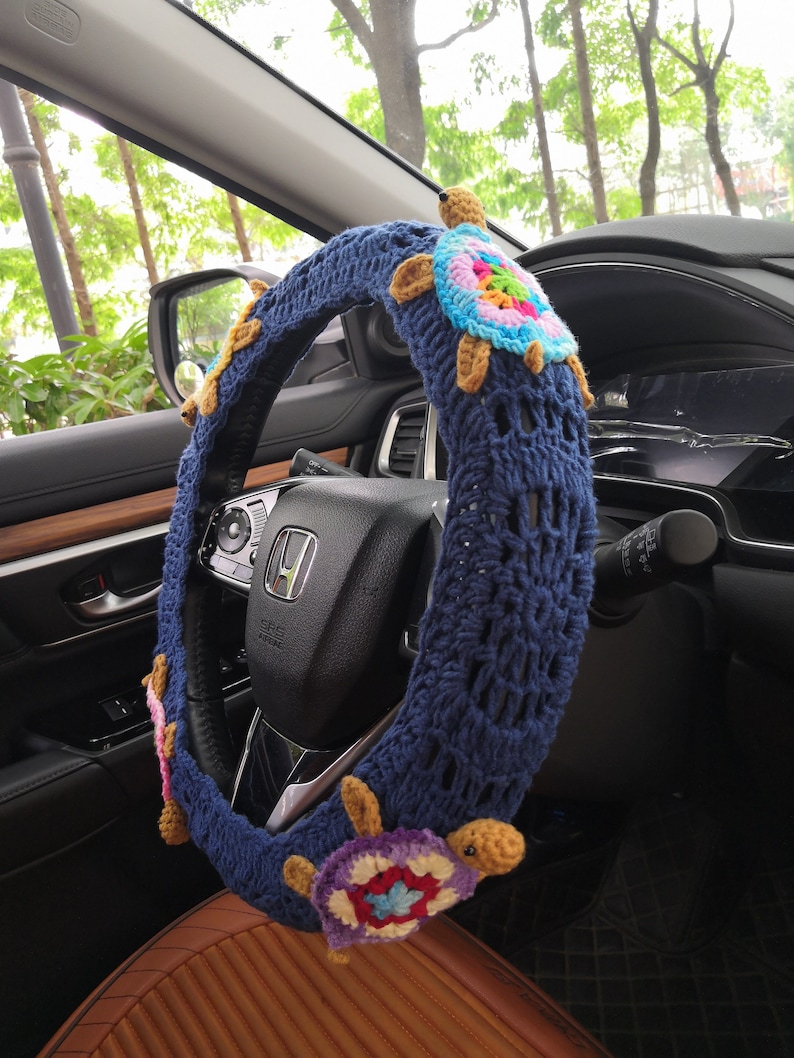 May include: A blue crocheted steering wheel cover with colorful crocheted turtle embellishments. The cover is on a car steering wheel.