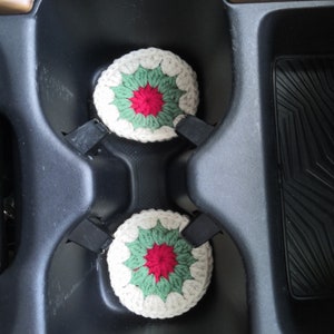 May include: Two crocheted cup coasters in a car cup holder. The coasters are white with green and red accents. The coasters are in the shape of a flower.