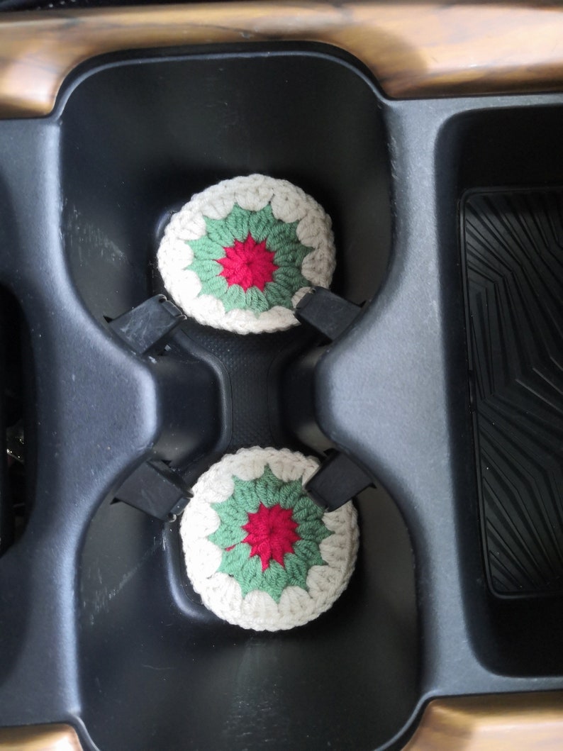 May include: Two crocheted cup coasters in a car cup holder. The coasters are white with green and red accents. The coasters are in the shape of a flower.
