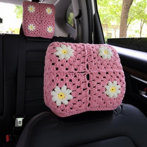 May include: Two pink crocheted headrest cushions with white daisy flowers. The cushions are in the backseat of a car.