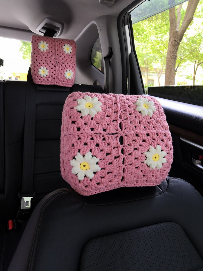May include: Two pink crocheted headrest cushions with white daisy flowers. The cushions are in the backseat of a car.