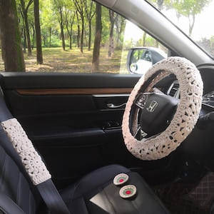 May include: A white crocheted steering wheel cover in a car interior. The car seat is black leather with a white crocheted seatbelt cover. Two small crocheted coasters with red and green embroidery are on the seat.