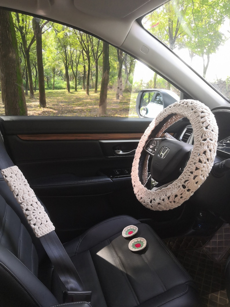 May include: A white crocheted steering wheel cover in a car interior. The car seat is black leather with a white crocheted seatbelt cover. Two small crocheted coasters with red and green embroidery are on the seat.