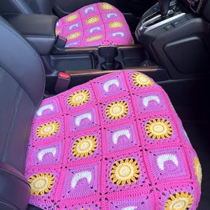 May include: Pink and yellow crochet car seat covers. The covers feature a square pattern with yellow sunflowers and white crescent shapes on a pink background. The covers are shown in a car interior.