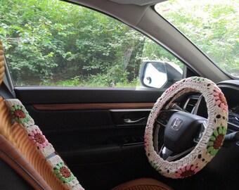 May include: A crocheted steering wheel cover with pink and green flowers. The cover is on a brown leather steering wheel in a car.