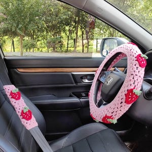 May include: Pink crocheted steering wheel cover with strawberry accents. The cover is on a black steering wheel in a car. There is a pink crocheted seatbelt cover with strawberry accents in the foreground.