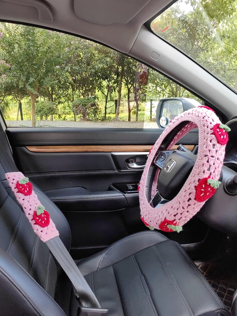 May include: Pink crocheted steering wheel cover with strawberry accents. The cover is on a black steering wheel in a car. There is a pink crocheted seatbelt cover with strawberry accents in the foreground.