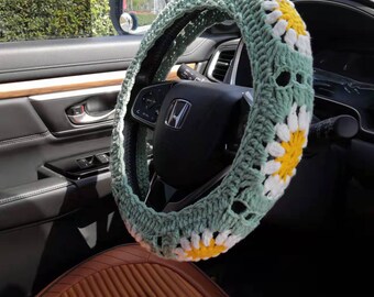May include: A green crocheted steering wheel cover with white and yellow daisy flowers. The cover is on a car steering wheel. Two matching daisy-shaped crocheted coasters are on the car seat.