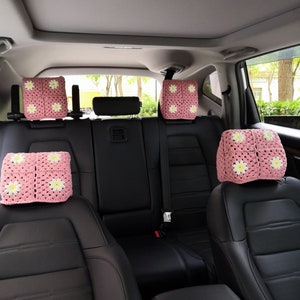 May include: Four pink crocheted headrest covers with white daisy accents are placed on the headrests of the back seats of a car.