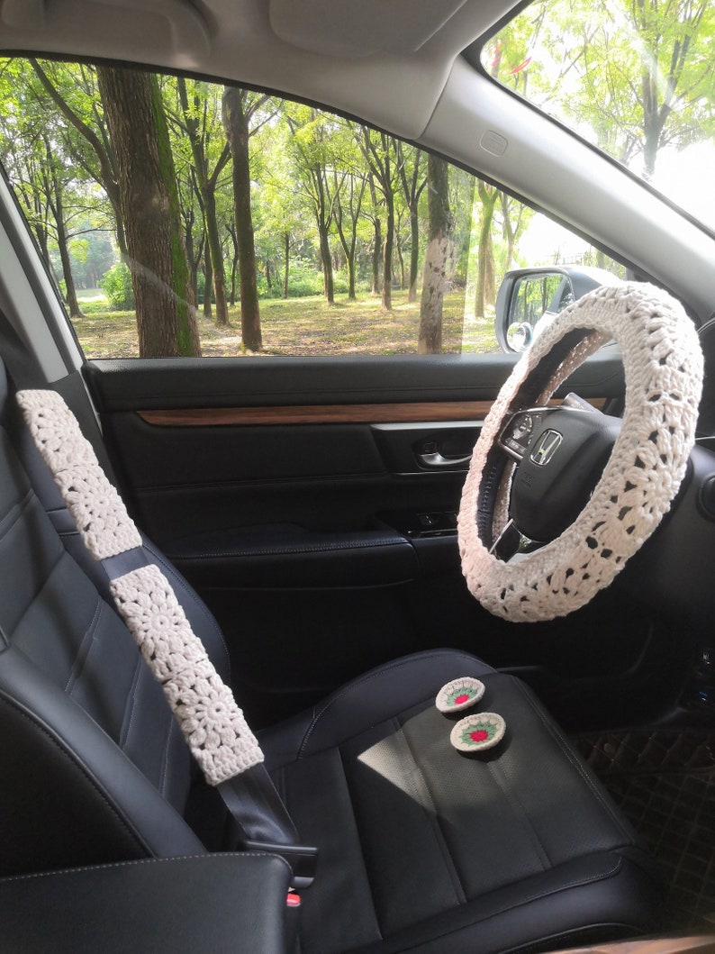 May include: A white crocheted steering wheel cover, seatbelt covers, and two crocheted coasters with red and green accents in a car interior.