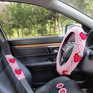 May include: Pink crocheted car accessories with a strawberry design. The set includes a steering wheel cover, seat belt covers, and two small seat cushions.