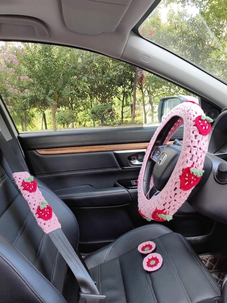 May include: Pink crocheted car accessories with a strawberry design. The set includes a steering wheel cover, seat belt covers, and two small seat cushions.