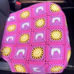 May include: A handmade crochet car seat cover featuring a vibrant patchwork design. The cover showcases a repeating pattern of yellow sunflowers, white arches, and purple and pink squares. The crochet work is visible, highlighting the texture and craftsmanship.