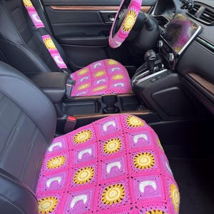 May include: Pink crochet car seat covers, steering wheel cover, and seat belt covers with a yellow sunflower and white crescent moon design. The interior of a car is visible, with black leather seats and a dashboard.