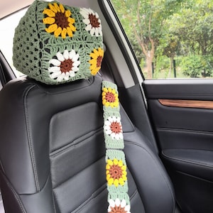 May include: Crocheted car headrest cover and seatbelt cover set. The headrest cover is olive green with sunflower motifs in yellow, brown, and white. The seatbelt cover has the same sunflower design. The set is placed on a black car seat.
