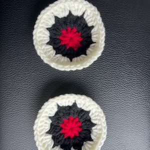 May include: Two circular crochet coasters featuring a central red star-like pattern surrounded by a black ring and finished with a thick white outer border. The yarn texture is visible throughout the pieces, creating a distinct layered design.