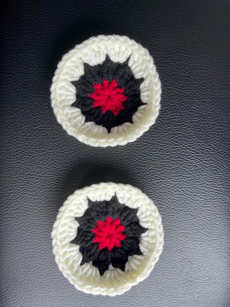 May include: Two circular crochet coasters featuring a central red star-like pattern surrounded by a black ring and finished with a thick white outer border. The yarn texture is visible throughout the pieces, creating a distinct layered design.