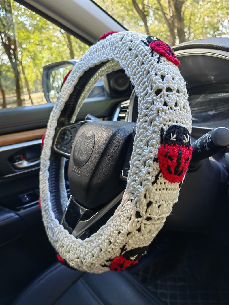 May include: A white crocheted steering wheel cover featuring multiple red and black ladybug appliques attached to the textured yarn surface. The cover is fitted over a black steering wheel inside a vehicle, showcasing a detailed stitch pattern.