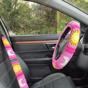 May include: Pink, yellow, and white crochet car accessories, including a steering wheel cover and seatbelt covers. The steering wheel cover features a sun design, while the seatbelt covers have sun and moon motifs. The car interior is black.