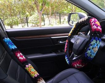 May include: A colorful crocheted steering wheel cover and seatbelt cover in a car interior. The cover is made of multi-colored yarn and features a square pattern.
