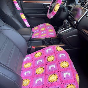 May include: A car interior featuring a crocheted steering wheel cover, seat covers, and seat belt covers. The crochet work is primarily pink, with yellow and white sunflower designs. The car's interior is black, with a digital display.