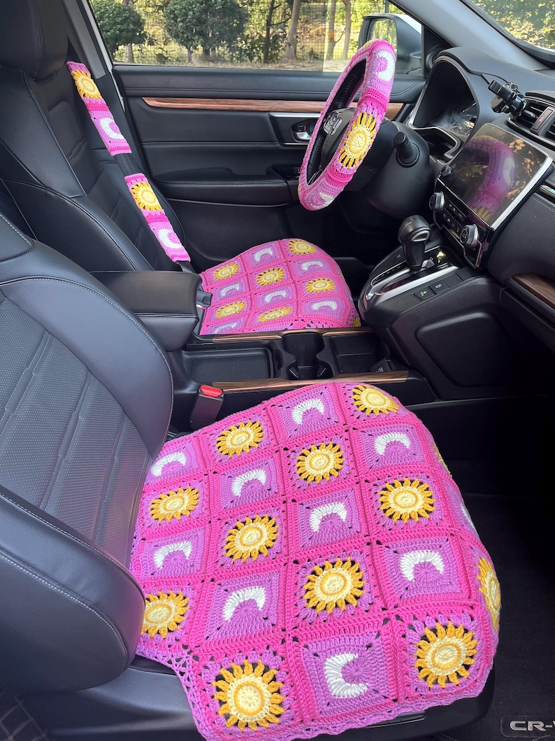 May include: A car interior featuring a crocheted steering wheel cover, seat covers, and seat belt covers. The crochet work is primarily pink, with yellow and white sunflower designs. The car's interior is black, with a digital display.