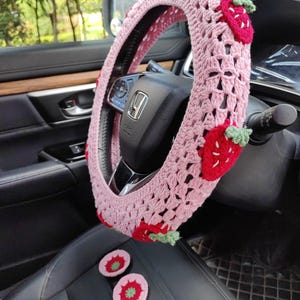 May include: Pink crocheted steering wheel cover with strawberry accents. The cover is made with a delicate crochet pattern and features embroidered strawberries with green leaves. Two additional crocheted strawberry accents are shown on the seat.