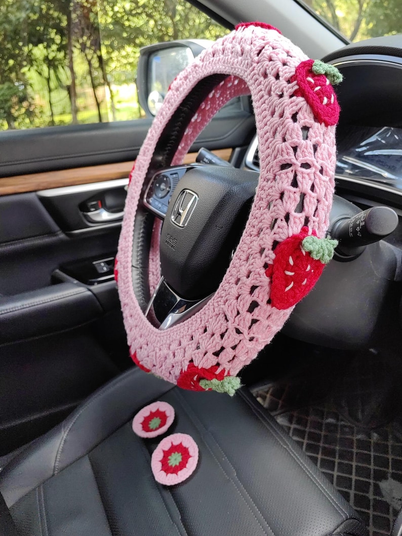 May include: Pink crocheted steering wheel cover with strawberry accents. The cover is made with a delicate crochet pattern and features embroidered strawberries with green leaves. Two additional crocheted strawberry accents are shown on the seat.