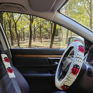 May include: A crochet steering wheel cover and a matching seat belt cover, both featuring a cream-colored base with a repeating pattern of red and black ladybug motifs. The accessories are installed inside a vehicle with black leather seats.