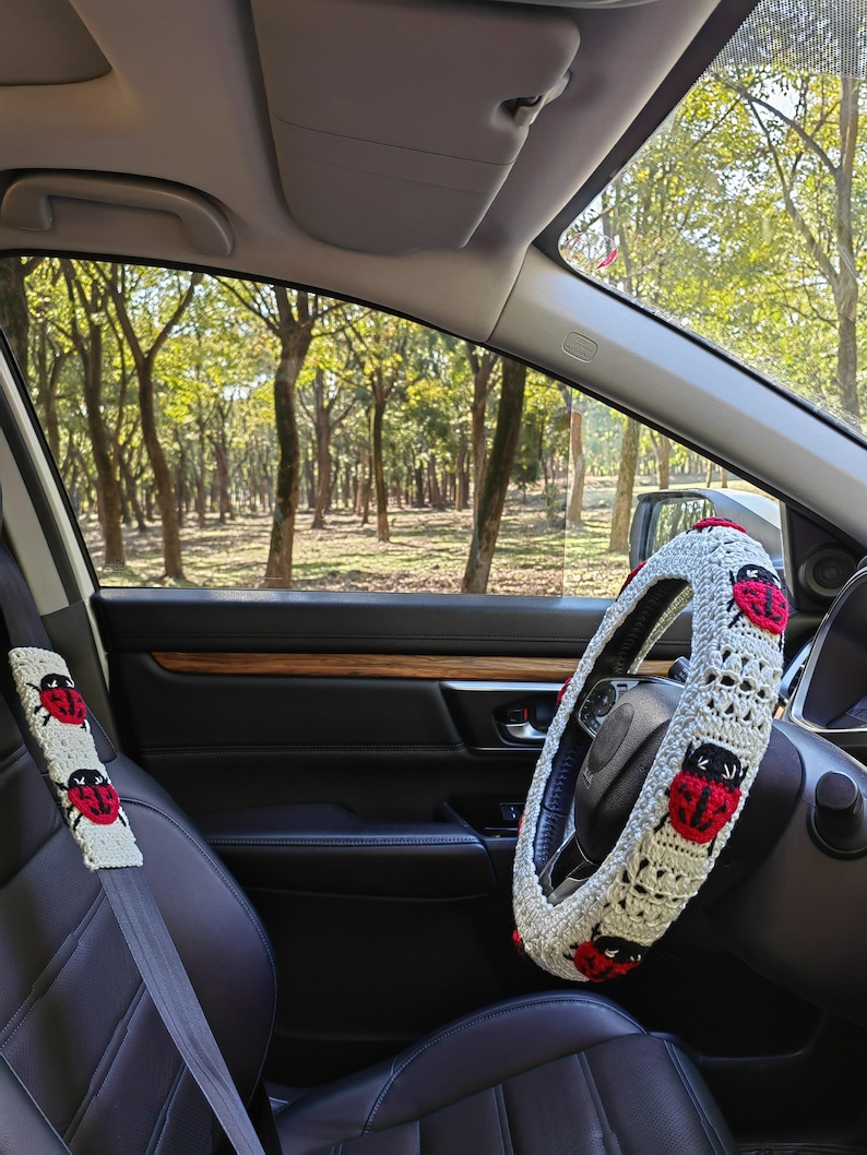 May include: A crochet steering wheel cover and a matching seat belt cover, both featuring a cream-colored base with a repeating pattern of red and black ladybug motifs. The accessories are installed inside a vehicle with black leather seats.