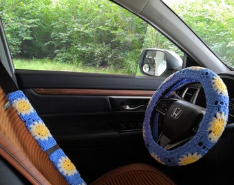May include: A blue crocheted steering wheel cover with white daisy flowers. The cover is installed on a black steering wheel with a silver Honda logo. The steering wheel is in a car interior with brown leather seats.