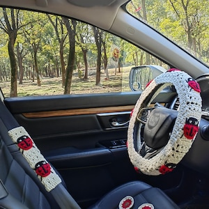 May include: A crochet steering wheel cover, a matching seat belt cover, and two small circular coasters, all featuring a cream-colored base with red and black ladybug motifs. The items are displayed inside a vehicle with black leather seats.