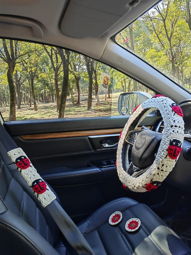May include: A crochet steering wheel cover, a matching seat belt cover, and two small circular coasters, all featuring a cream-colored base with red and black ladybug motifs. The items are displayed inside a vehicle with black leather seats.
