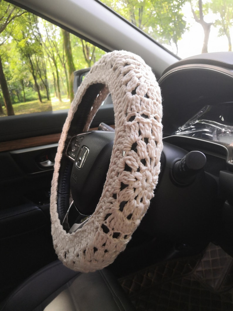 May include: A close-up of a beige crocheted steering wheel cover. The cover is made of a delicate, open-weave pattern and is designed to fit over a standard steering wheel.