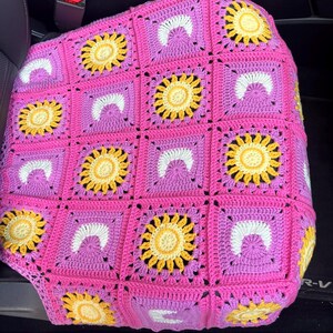 May include: A crocheted seat cover featuring a vibrant pink background with square patterns. Each square showcases a yellow sunflower design, a white crescent, and a purple center. The handmade cover is placed on a car seat.