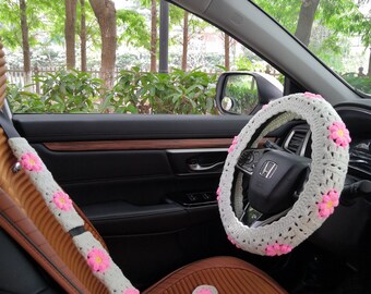 May include: White crocheted steering wheel cover with pink flower accents. The cover is installed on a car's steering wheel. The car's interior is brown and black. There are also matching pink flower seatbelt covers and seat cushions.