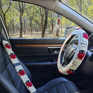 May include: A cream-colored crochet steering wheel cover and a matching seat belt cover, both featuring a repeating pattern of red and black ladybug motifs. The accessories are installed on a black leather steering wheel and seat belt.
