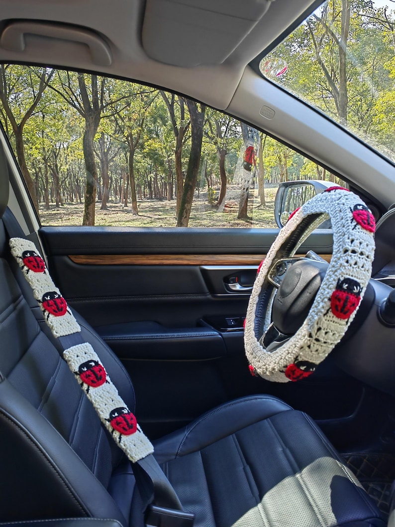 May include: A cream-colored crochet steering wheel cover and a matching seat belt cover, both featuring a repeating pattern of red and black ladybug motifs. The accessories are installed on a black leather steering wheel and seat belt.
