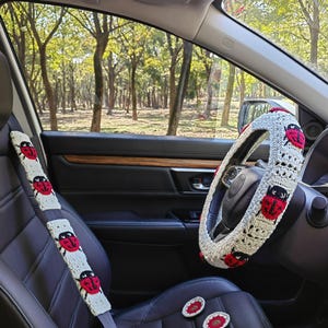 May include: A cream-colored crochet steering wheel cover and a matching seat belt cover, both featuring red and black ladybug motifs. Two small, round, matching crochet coasters are placed on the black leather car seat.