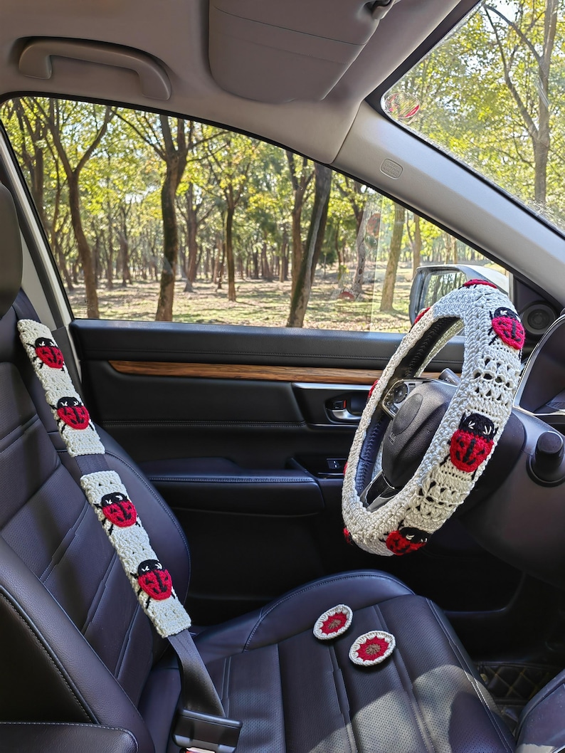 May include: A cream-colored crochet steering wheel cover and a matching seat belt cover, both featuring red and black ladybug motifs. Two small, round, matching crochet coasters are placed on the black leather car seat.
