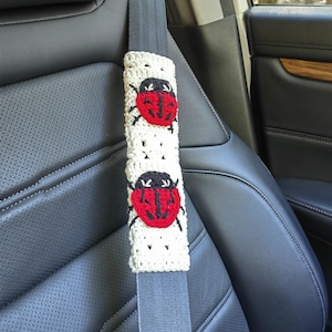May include: A cream-colored crochet seat belt cover featuring two red and black ladybug appliqu&eacute;s. The rectangular cover is wrapped around a gray seat belt strap, showcasing a textured, open-stitch pattern throughout the fabric.