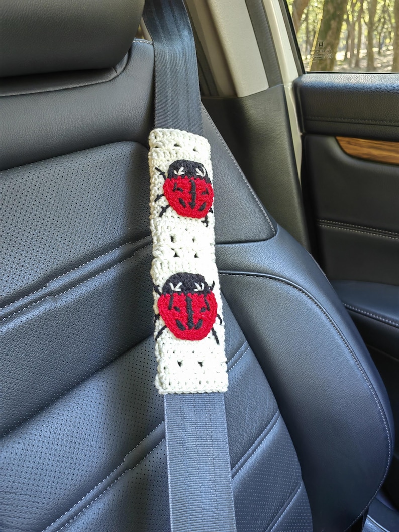 May include: A cream-colored crochet seat belt cover featuring two red and black ladybug appliqu&eacute;s. The rectangular cover is wrapped around a gray seat belt strap, showcasing a textured, open-stitch pattern throughout the fabric.