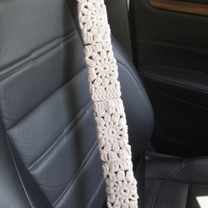 May include: A white crocheted car seatbelt cover with a floral design. The cover is attached to a black seatbelt and is hanging in a car.