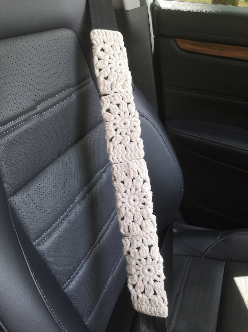 May include: A white crocheted car seatbelt cover with a floral design. The cover is attached to a black seatbelt and is hanging in a car.