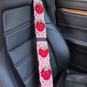 May include: Pink crocheted car seatbelt cover with embroidered strawberries. The cover is made of pink yarn and features four red strawberries with green leaves.