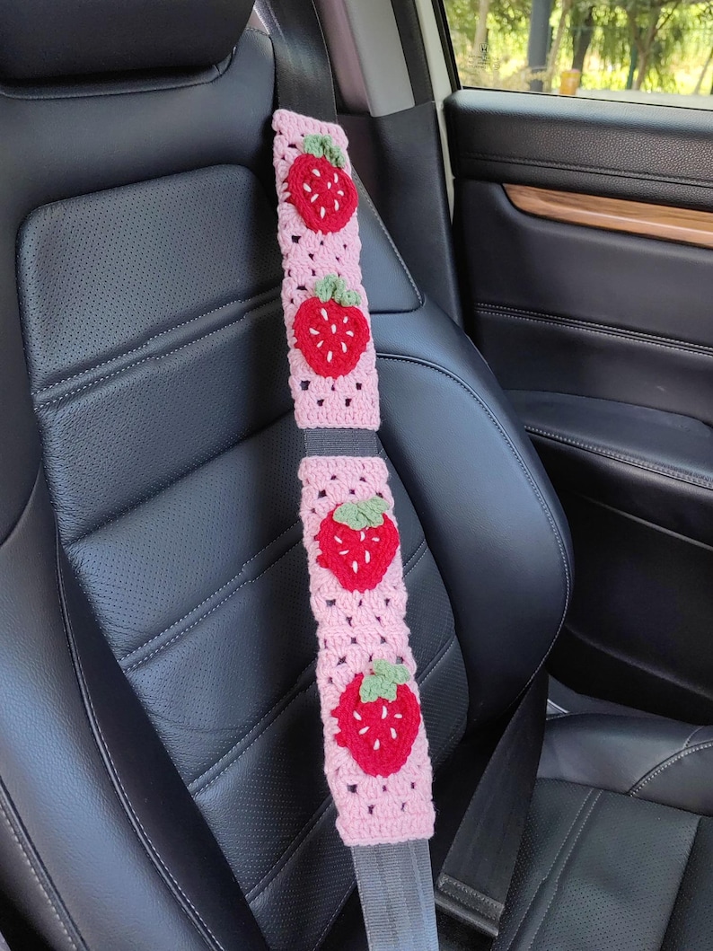 May include: Pink crocheted car seatbelt cover with embroidered strawberries. The cover is made of pink yarn and features four red strawberries with green leaves.
