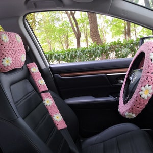 May include: Pink crocheted car seat covers with white daisy accents. The set includes a headrest cover, two seat belt covers, and a steering wheel cover.