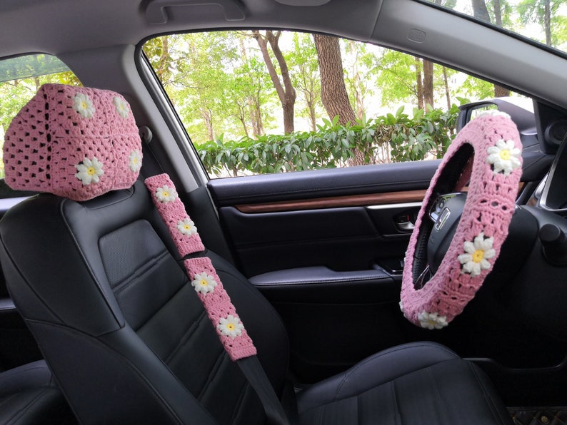 May include: Pink crocheted car seat covers with white daisy accents. The set includes a headrest cover, two seat belt covers, and a steering wheel cover.